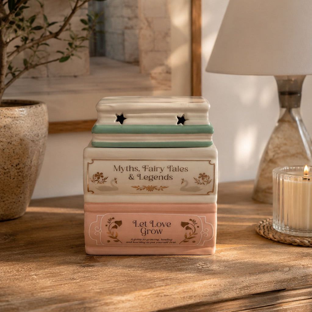 Quemador de cerámica en forma de libros apilados en tonos blanco, verde y rosa sobre una mesa de madera