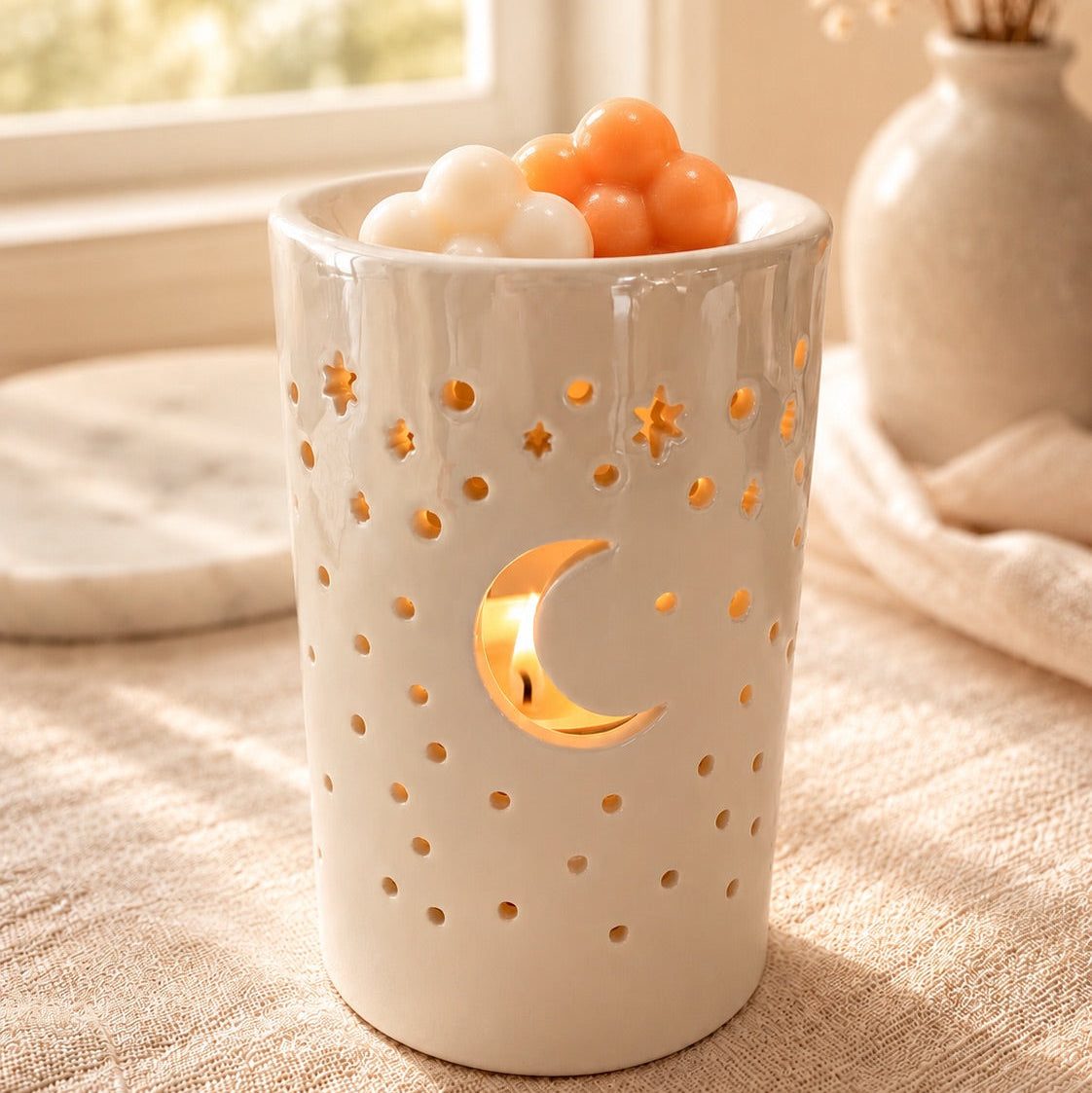 Quemador Luna de cerámica blanca con calados de luna y estrellas, encendido y con wax melts en forma de nubes en el plato superior.